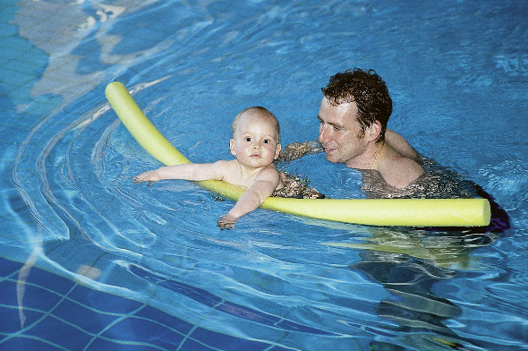 Baby schwimmt mit einer Schwimmnudel im Wasser und der Vater schwimmt hinteher